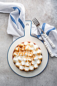 View from above of a beautiful homemade lemon cake on a round plate. A striped napkin and cutlery lie next to it, against a textured background.