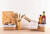 A modern kitchen with eco-friendly bamboo chopping board, utensils and storage containers, as well as jars of spices and bottles of cooking oils, arranged on a wooden worktop against a backdrop of white bricks.