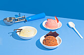 Three flavours of ice cream, vanilla, strawberry and chocolate, served in white bowls on a bright blue background. A scoop and a white spoon complete the scene.