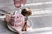 An artistic Easter table arrangement with a pink fabric cloth with a bunny motif, elegantly tied, accompanied by unique pink glasses and a delicate willow nest with speckled eggs, all set against a backdrop of soft light and shadow.