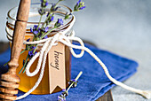 A jar of golden honey, decorated with a ribbon of twine, small sprigs of lavender and a handwritten label A wooden honey spoon rests next to it on a blue fabric background