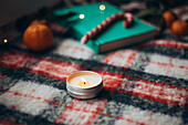 Warm Christmas scene with a lit candle, scattered mandarins, a candy cane and books on a festive chequered blanket.