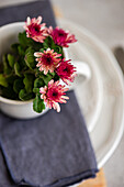 Vibrant pink chrysanthemum flowers in a white cup on a layered, textured background of grey fabric and white plate create a cheerful and elegant table setting