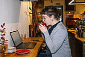 A woman enjoys a cup of speciality coffee while working on her laptop in a cosy, modern café. Productivity and relaxation merge seamlessly in the inviting ambience.