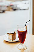A glass of iced tea with a straw and a slice of cake on a wooden table near a window, offering a relaxed atmosphere in a cosy café