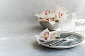 A calmly laid table with delicate orchids on a pastel-coloured plate, accompanied by silver cutlery and textured glasses that convey a modern and elegant dining room ambience.