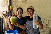 Two smiling Asian women present a freshly opened coconut and a bottle of coconut water, emphasising their refreshing taste. A perfect moment in a rustic setting.