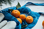 Fresh mandarins are presented in a bright blue bag decorated with twinkling fairy lights next to festive Christmas decorations.