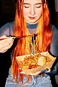 A woman with long orange hair enjoys a noodle dish with chopsticks at an Asian food market The scene captures the vibrancy and diversity of street food culture