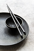 Minimalist black placemat with a textured plate, a small bowl and detailed chopsticks. The monochrome design creates a simple and sophisticated aesthetic for dining.