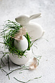 A carefully designed Easter table with a white egg in bright green grass, accentuated with delicate feathers, and an elegant ceramic bunny on a textured surface.