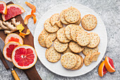 A white plate with homemade ginger and grapefruit biscuits, some of which are broken, against a grey background. Slices of fresh pink grapefruit and ginger root lie on a wooden chopping board. Citrus peel is scattered across the board. A knife with a wooden handle lies on the worktop next to the plate. This image conveys a sense of homemade cosiness and the delicious taste of citrus and ginger.