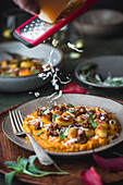 A side view of a plate of gnocchi and sausage with sage and parmesan on a pumpkin sauce. One hand holds a red grater with which Parmesan cheese is sprinkled over the dish. A rustic, autumnal table setting can be seen in the background.