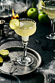Lime Margarita with salt rim on black surface, Halloween setting, lime slices on black tray