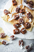 A close-up of homemade caramel sweets on a marble surface. The candies are wrapped in white parchment paper and sprinkled with sea salt. They are arranged on a piece of brown parchment paper standing on a white marble cake stand. More caramels can be seen in the blurred background, hinting at a delicious treat and highlighting the simplicity and beauty of homemade sweets.