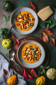 A wide-angle shot of two bowls of crispy gnocchi in pumpkin sauce with sage and sausage on a dark green surface. The bowls are surrounded by autumn decorations, including pumpkins, leaves and a sprig of sage. The cutlery lies on a wooden tray, and on the other side is a small plate with a piece of Parmesan cheese. The picture is a beautiful and appetising depiction of autumn flavours.