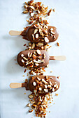 Three almond ice cream sticks covered in milk chocolate, sprinkled with roasted almonds on a white marble base