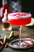 Close-up of a sophisticated alcoholic drink, with a textured, stemmed coupe cocktail glass with colourful red liqueur topped with foam, rhubarb and pink berry powder as garnish