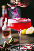 An action shot of bright pink berry powder sprinkled on a sophisticated alcoholic drink with a textured, stemmed coupe cocktail glass with colourful red liqueur and a frothy top