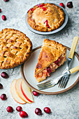 A close-up of 2 freshly baked mini apple and cranberry tarts. One piece is on a small plate. Apple slices and cranberries are scattered on the plate. The tart is golden brown and the filling is visible through the crust.