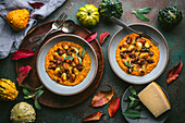 Wide-angle shot of two bowls of crispy gnocchi in pumpkin sauce with sage and sausage on a dark green surface. The bowls are surrounded by autumn decorations, including pumpkins, leaves and a sprig of sage. The cutlery lies on a wooden tray, and on the other side is a small plate with a piece of Parmesan cheese. The picture is a beautiful and appetising depiction of autumn flavours.