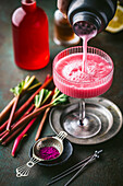 Action shot of a pink rhubarb cocktail being poured from the shaker into a beautiful half-full coupe glass with fresh rhubarb and red syrup in the background
