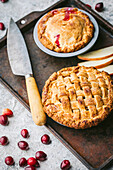 Two mini apple pies, both with a golden brown crust, lie on a rustic, weathered baking tray on a grey worktop. One pie has a lattice lid, the other a solid lid through which a hint of red cranberry filling shines through. A few apple slices and an old kitchen knife lie next to the cakes. The picture conveys a feeling of warmth and cosiness, as if the cakes have just come out of the oven.