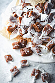 A close-up of a large stack of homemade caramels on a marble surface. The sweets are wrapped in white parchment paper and sprinkled with sea salt. They are arranged on a piece of brown parchment paper standing on a white marble cake stand. More caramels can be seen in the blurred background, hinting at a delicious treat and highlighting the simplicity and beauty of homemade sweets.