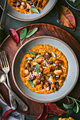 Close-up of a bowl of crispy gnocchi in pumpkin sauce with sage and sausage on a dark green surface. Surrounded by autumn decorations, including pumpkins, leaves and a sprig of sage. A wooden tray holds the silver cutlery. The picture is a beautiful and appetising depiction of autumn flavours.
