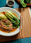 Bowl of soba noodles in spicy broth with baby bok choy, surrounded by fresh mushrooms, ginger