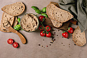 Homemade liver pâté in a glass bowl with slices of wholemeal bread, fresh cherry tomatoes and basil leaves on a wooden chopping board. Rustic breakfast setting on a beige background.