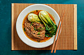Bowl of soba noodles in a spicy broth with baby bok choy, surrounded by fresh mushrooms, ginger