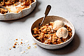 A bowl of apple crumble with two scoops of vanilla ice cream, dusted with cinnamon, and a partially eaten bowl in the background.