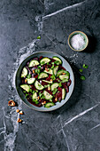 Vegan beetroot salad. Marinated, sliced beetroot with fresh cucumber, walnuts and coriander leaves in a ceramic bowl. Next to it a bowl with sea salt on a black marble table. Healthy, plant-based dish. View from above