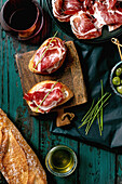 Sliced Spanish ham Jamon Iberico paleta on a black plate. Bowl of green olives, fresh and toasted white bread, olive oil, glass of red wine, tapas. Old wooden background, dark turquoise colour. Place to copy