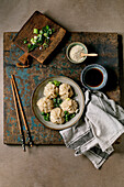 Homemade Asian steamed dumplings in a grey plate, topped with sesame seeds and green onions. Soy sauce, chopsticks, grey napkin nearby on a rustic wooden board, beige textured background.