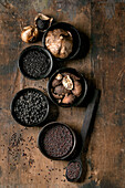 Set of five black spices: black fermented garlic, black Himalayan salt, black mustard seeds, black cardamom and fermented campota pepper in black ceramic bowls. Dark wooden background. Close-up