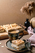 Homemade Norwegian cake Verdens Beste Kake with pudding, meringue and almonds, cut into squares, served on a wooden tray. Beige-coloured table with coffee cup, spoon, milk jug, dry reed in a bottle, beige-coloured wall.