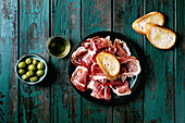 Thinly sliced Spanish Jamon Iberico ham on a black plate. Bowl with green olives, toasted white bread, olive oil. Old wooden background, dark turquoise colour, view from above.