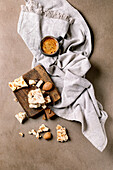 Traditional Spanish dessert: crispy turron with almonds, broken into pieces and served on a wooden board. Linen napkin, espresso coffee in black cup. Beige-coloured textured background, view from above.