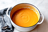 A pot of creamy carrot soup sits on a light-coloured surface with a grey linen cloth and a silver spoon next to it.