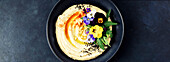 A beautifully presented bowl of creamy hummus, garnished with olive oil, black sesame seeds, pine nuts, capsicum, rocket and vibrant edible flowers, served with flatbread on a dark textured background. Perfect as a banner image for food-related content.