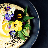 A close-up of creamy hummus, garnished with olive oil, black sesame seeds, rocket and vibrant edible flowers, served on a dark plate. A luxurious and artistic interpretation of a Mediterranean classic.