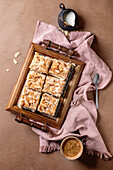 Homemade Norwegian sweet cake Verdens Beste Kake with pudding, meringue and almonds, cut into squares, served on a wooden tray. Beige background, pink fabric, coffee cup, spoon, milk jug. View from above