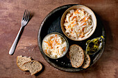 Traditional starter Sauerkraut with carrots in a bowl and glass. Sea salt, olive oil, sliced rye bread and fork nearby. Healthy food, fermented products. Brown textured background, top view.