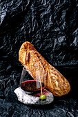 Still life with a glass of red wine and a single olive standing on a stone and a loaf of white bread. Crumpled black paper in the background. Concept of bread and wine.