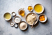 An assortment of baking ingredients, including eggs, sugar, lemon zest and flour, arranged on a textured surface.