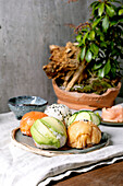 Temari sushi. Trendy sushi balls with salmon, avocado, cucumber and Japanese omelette on a wabi-sabi plate. Soy sauce, pickled ginger, ikebana on table with grey linen cloth. Grey textured background.