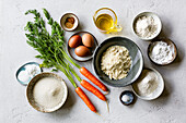An assortment of baking ingredients, including carrots, eggs, flour, sugar, spices, oil and baking powder, arranged on a white surface.