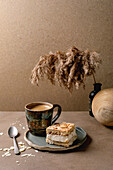 Homemade Norwegian sweet cake Verdens Beste Kake with vanilla pudding, meringue and almonds, a square piece on a plate on a beige table with coffee cup, spoon, milk jug, reed in bottle, beige wall.